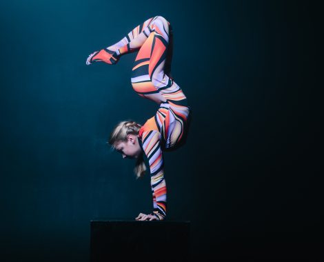 Young flexible blonde circus acrobat posing in studio in costume. Doing equilibre balance handstand on a cube. Copy space text.